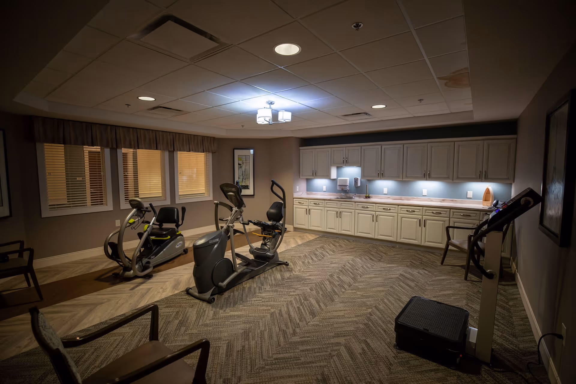 A small fitness room with exercise equipment including two recumbent bikes and a treadmill. The room has beige walls, a patterned carpet, overhead lighting, and a long counter with cabinets and a sink along one wall. There are also a few chairs placed around the room.