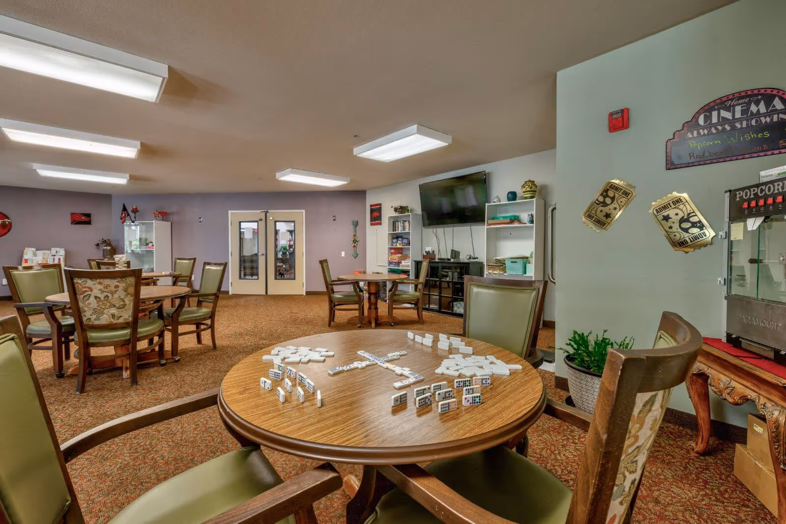 Community activity room with round tables and chairs, a table set up for dominoes, a wall-mounted TV and a popcorn machine.