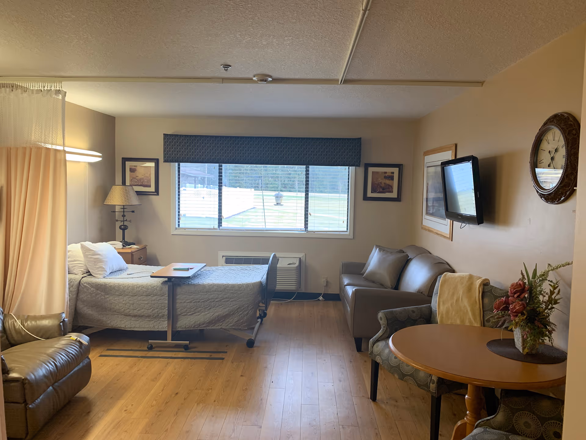 A well-lit room in a senior living facility featuring a hospital-style bed with white pillows and a beige quilt, a bedside table with a lamp, a brown leather recliner, a small round table with two patterned chairs and a floral arrangement, a gray couch, a wall-mounted flat screen TV, a large window with blinds, and a wall clock.