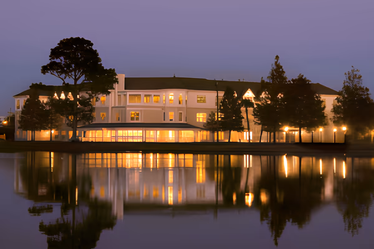 A large, well-lit building at dusk with multiple windows glowing warmly. The building is reflected in a calm body of water in the foreground, surrounded by trees.