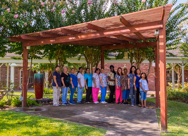 A group of people standing in a line under a wooden pergola in an outdoor garden area with trees and a brick building in the background.