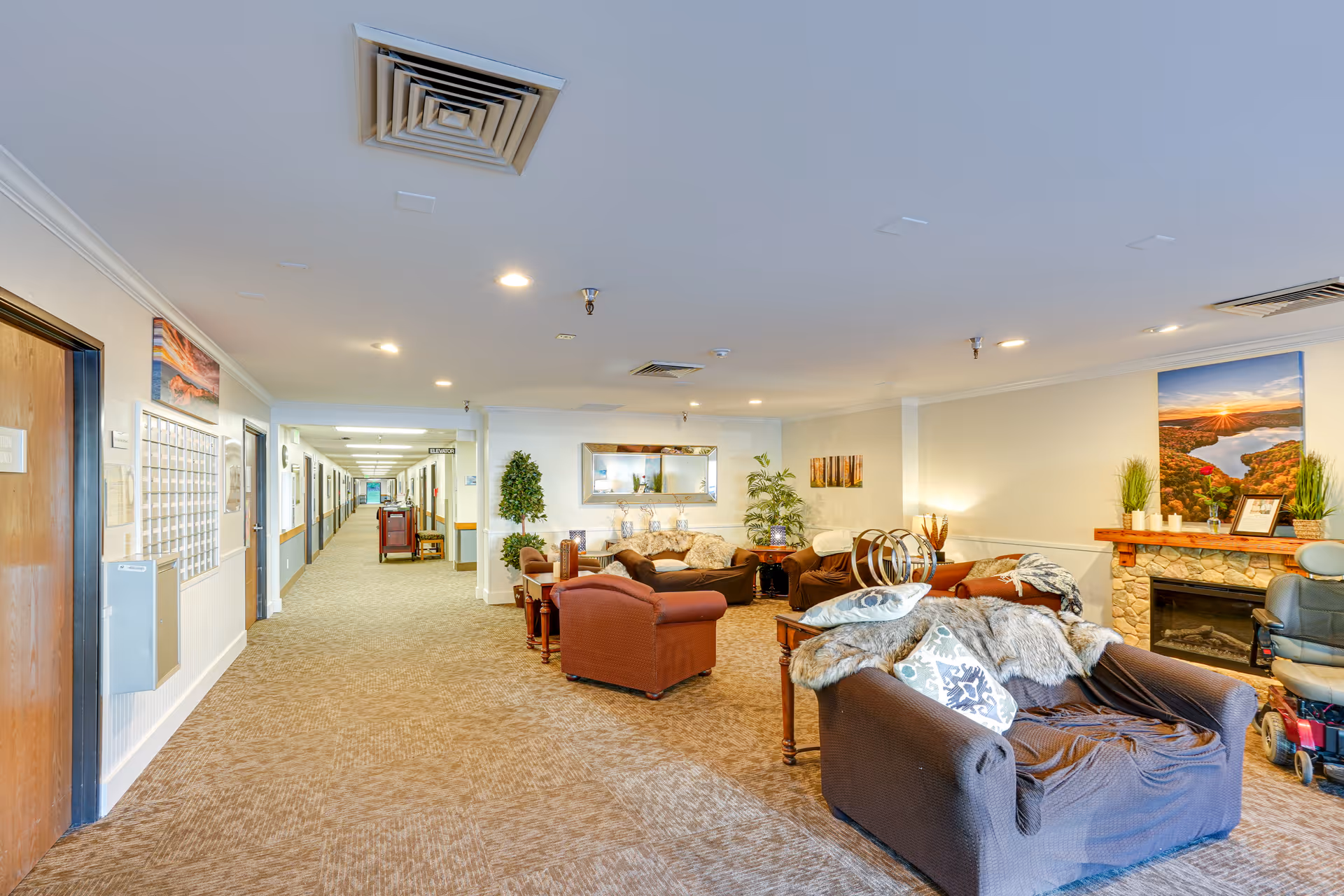 Spacious senior living common area with upholstered chairs and sofas, a fireplace, plants, and a long hallway of doors.