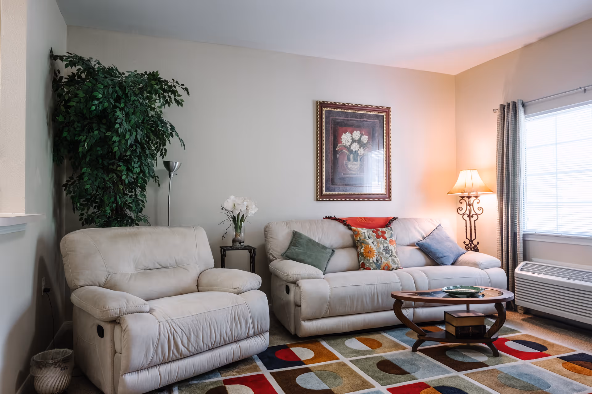 A cozy living room with a beige loveseat and sofa adorned with colorful throw pillows. A wooden coffee table sits on a multicolored geometric rug. There is a tall green plant in the corner, a side table with white flowers, a decorative floor lamp, and a window with curtains letting in natural light.