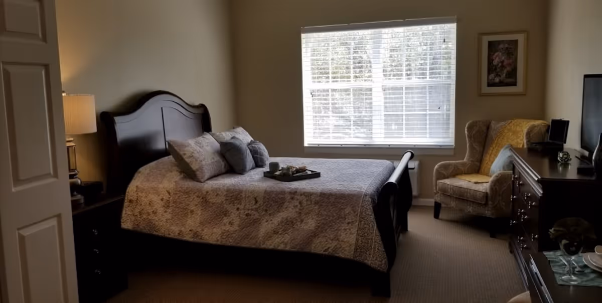 A cozy bedroom with a dark wooden bed frame, a neatly made bed with a patterned quilt and several pillows. A tray with a cup and other items is placed on the bed. To the left, there is a nightstand with a lamp. A large window with blinds lets in natural light. On the right side, there is a comfortable armchair with a yellow throw blanket and a framed picture on the wall above it. A dark wooden dresser with a TV on top is also visible.