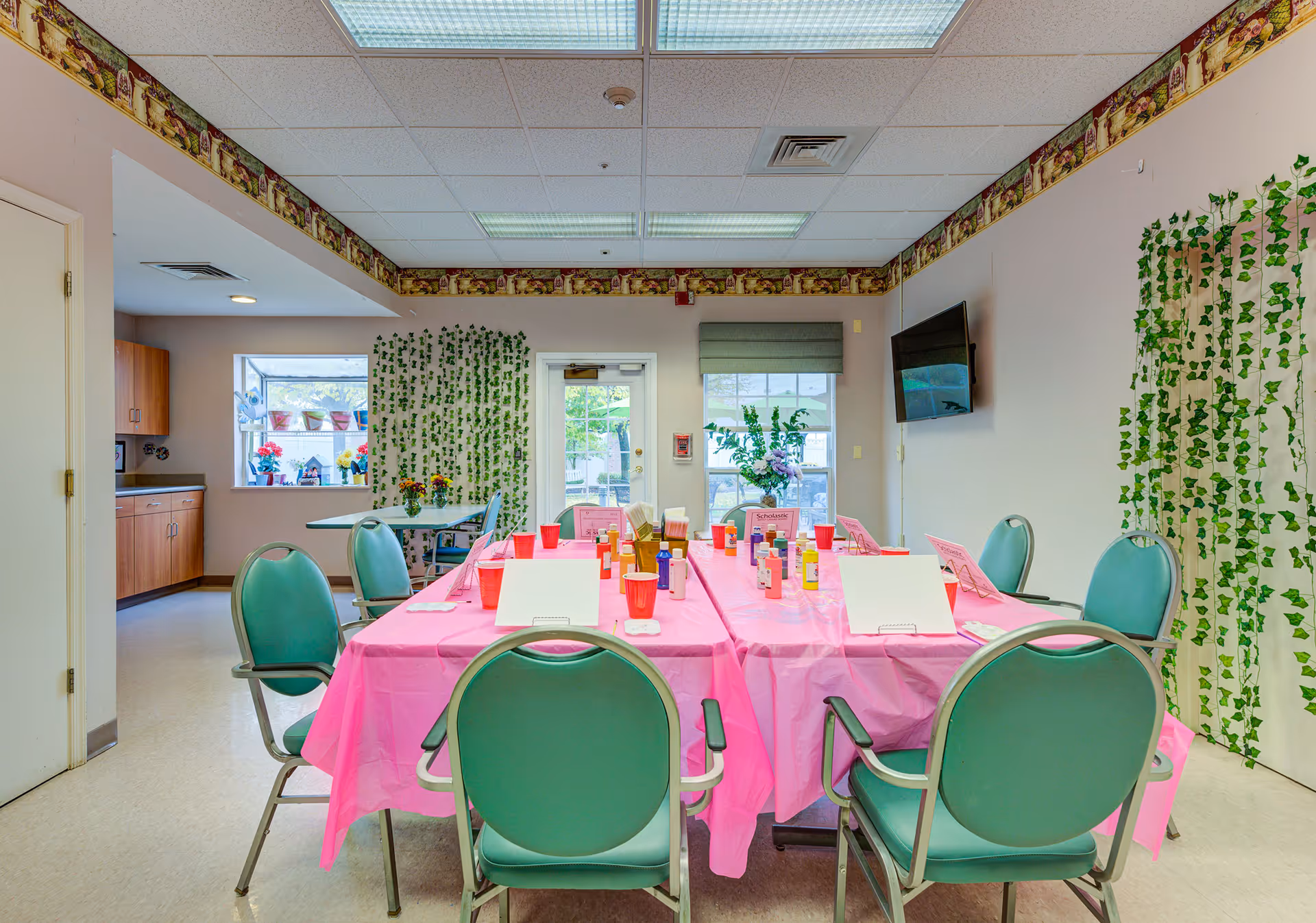 A brightly lit room with a table covered in a pink tablecloth set up for an arts and crafts activity. The table has green chairs around it, with various bottles of paint, red plastic cups, and white paper or canvas boards. The room has light-colored walls decorated with green leafy vines and a border near the ceiling. There is a window and a door at the back, a wall-mounted TV on the right, and a small kitchenette area visible on the left.