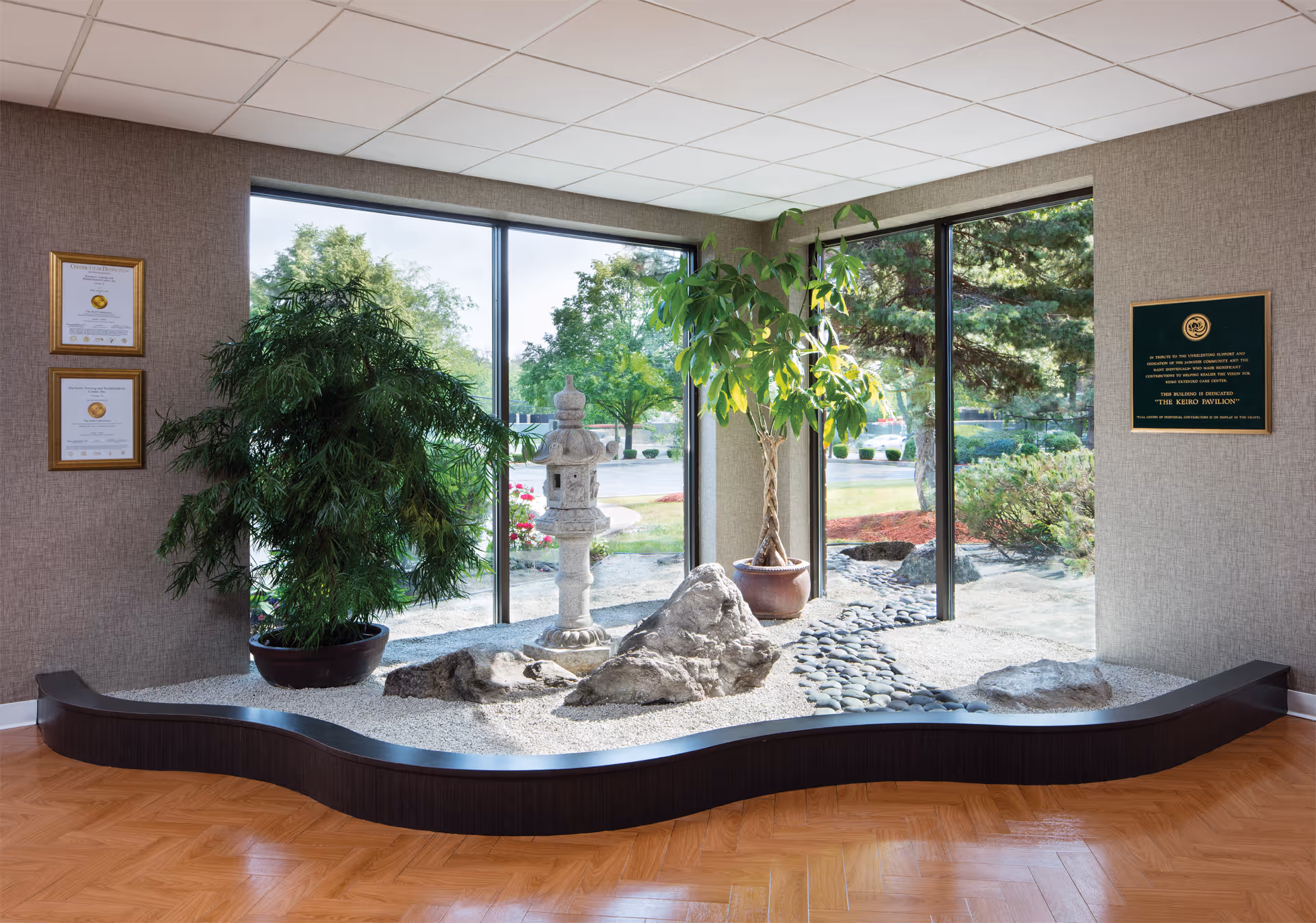 Indoor Japanese-style rock garden with large windows showing outdoor greenery. The garden features white gravel, large rocks, a stone lantern, and two potted plants. The room has wood flooring and textured walls with framed certificates and a plaque.