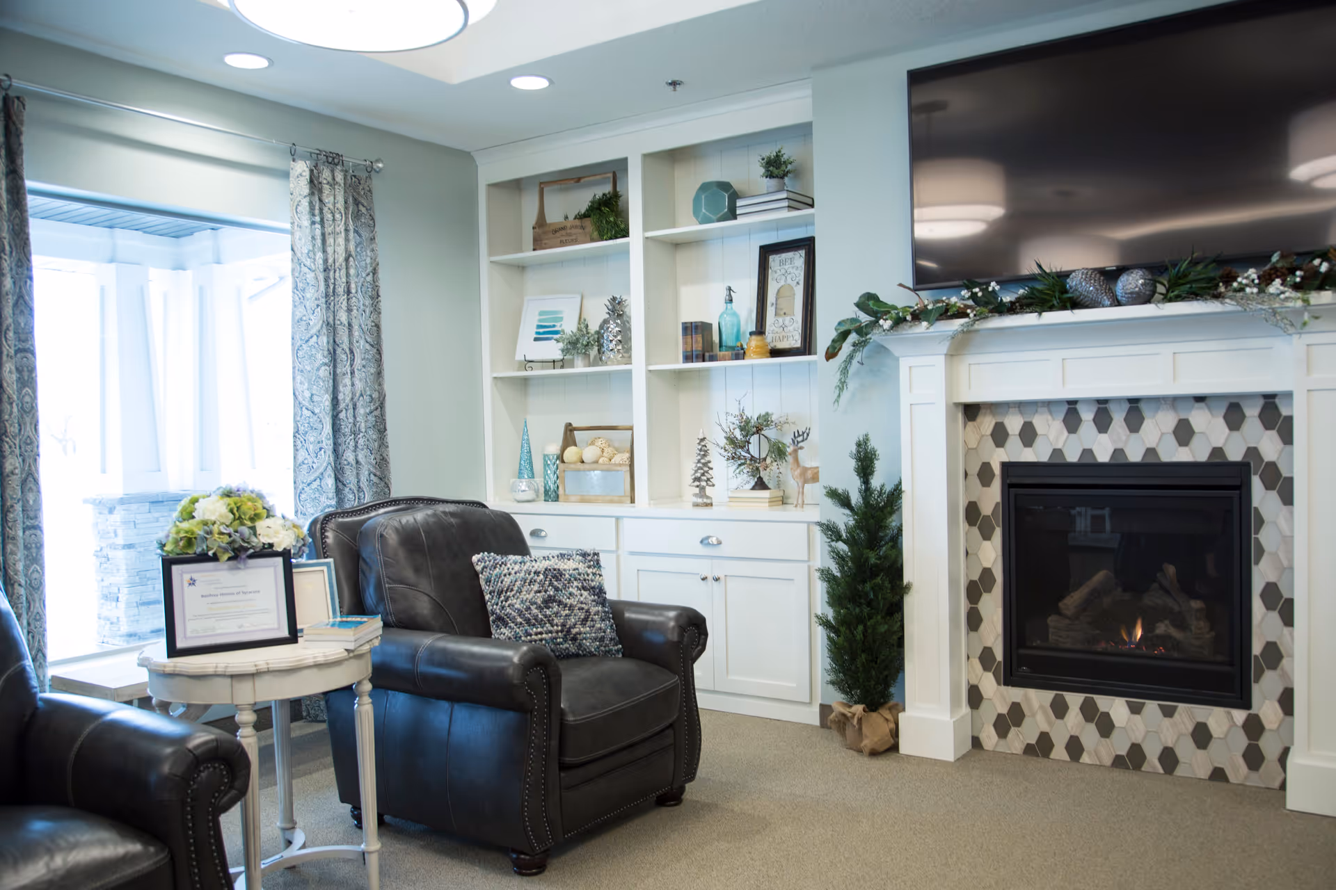 A cozy living room area with two black leather armchairs, a small round white side table with a flower arrangement and framed certificate, built-in white shelves decorated with various ornaments, and a fireplace with a hexagonal tile surround and a large flat-screen TV mounted above it.