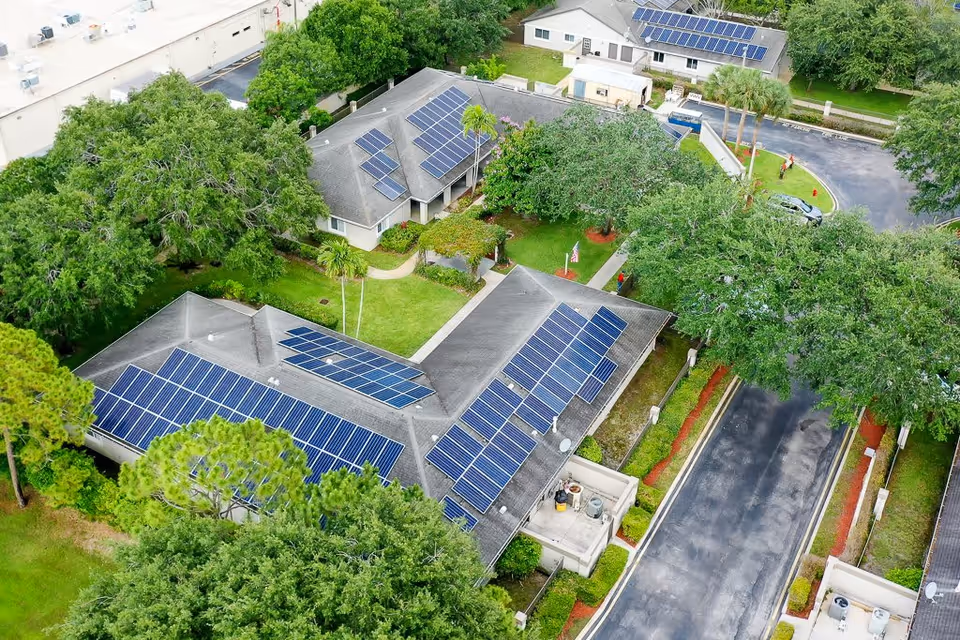 Aerial view of a senior living facility named Cottages at Palm Beach showing multiple single-story buildings with solar panels on the roofs, surrounded by trees, greenery, and paved roads.