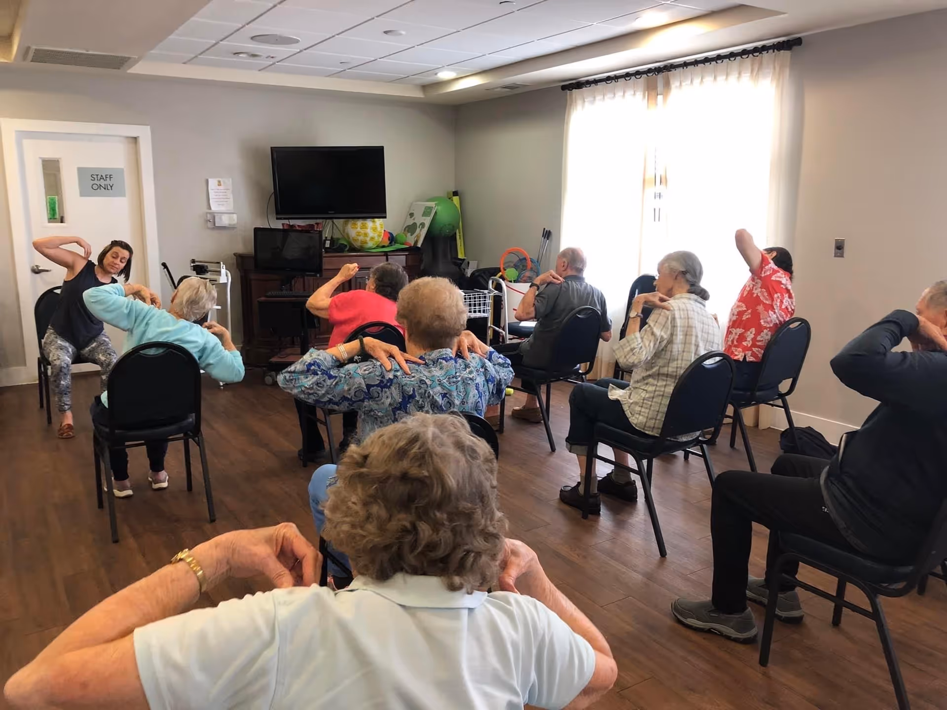 A group of elderly people seated on chairs in a room participating in a seated exercise class led by an instructor. The participants are stretching their arms behind their heads. The room has wooden flooring, a television mounted on the wall, and a window with curtains allowing natural light.