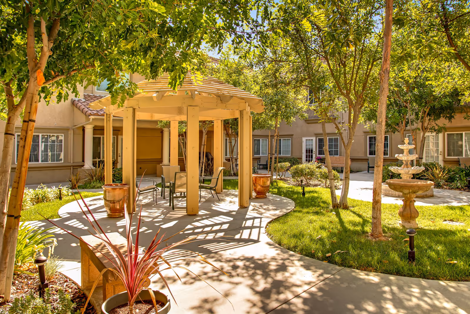 Outdoor courtyard area at Fairwinds - West Hills featuring a shaded pergola with chairs and tables, surrounded by green grass, trees, potted plants, and a multi-tiered stone fountain. The courtyard is enclosed by a beige building with multiple windows and doors.