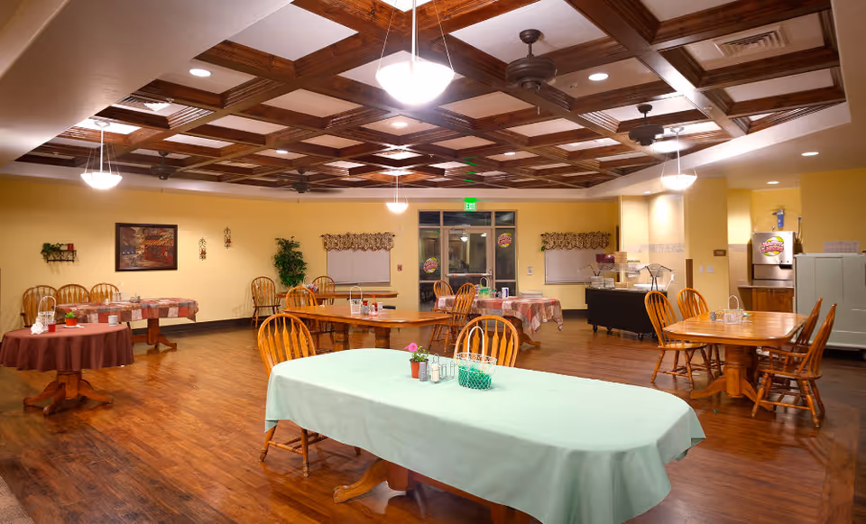 Spacious dining room with multiple wooden tables and chairs, a coffered ceiling with hanging lights, and a buffet area at the far wall.