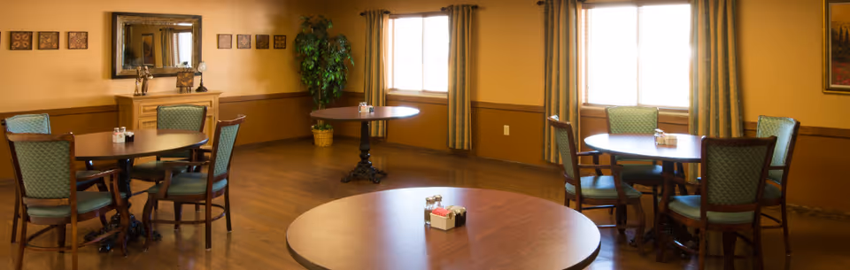 A dining room with several round wooden tables and green cushioned chairs arranged around them. The room has wooden flooring, beige walls with a wooden trim, two windows with curtains, a large mirror on one wall, decorative framed art pieces, and a potted plant in the corner.