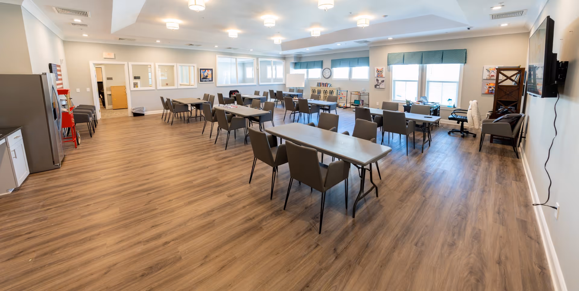 A spacious, well-lit room with multiple tables and chairs arranged in rows, suitable for group activities or dining. The room has wood flooring, several windows with blue valances, a wall-mounted TV, and various decorations including framed pictures and a clock. There is a kitchenette area with a refrigerator and a popcorn machine on the left side.