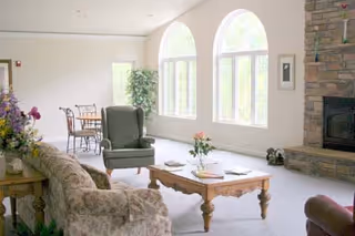 A bright living room with large arched windows letting in natural light. The room features a floral patterned sofa, a green armchair, a wooden coffee table with flowers and books, and a stone fireplace on the right side. In the background, there is a small dining table with chairs and a potted plant near the windows.