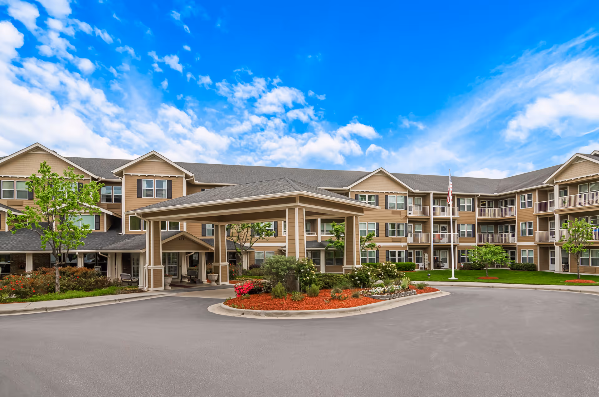 Front exterior view of a three-story senior living facility building with a covered entrance, landscaped garden beds with flowers and shrubs, and an American flag on a flagpole under a partly cloudy blue sky.