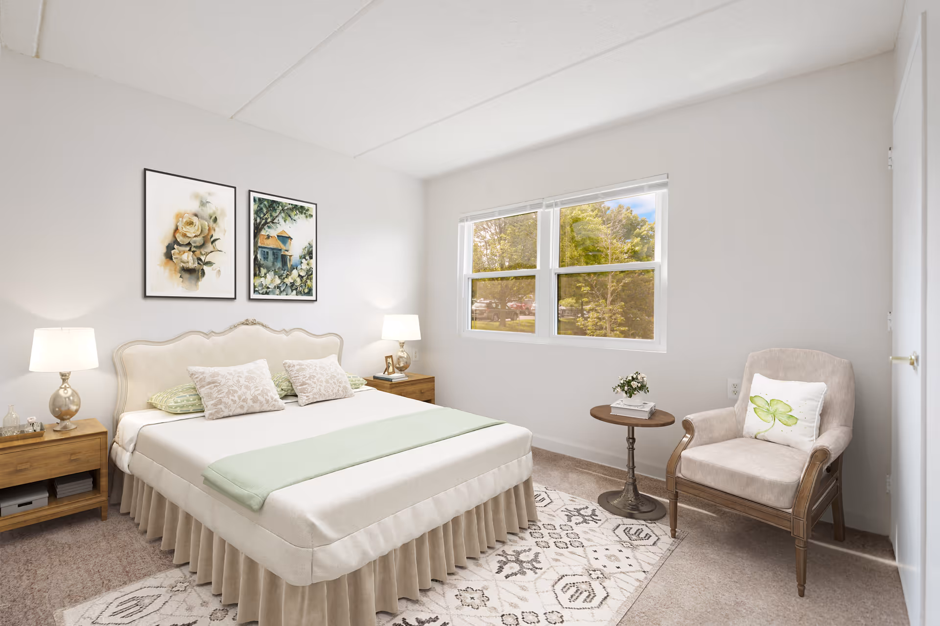 A bright and cozy bedroom with a large bed featuring a beige upholstered headboard, white bedding, and green accent pillows and throw. There are two wooden nightstands with lamps on either side of the bed. Above the bed, two framed floral and nature-themed paintings hang on the wall. A window lets in natural light, showing trees outside. A beige armchair with a green leaf-patterned pillow and a small round wooden side table with a book and a small plant are positioned near the window. The room has light-colored walls and carpeted floor with a patterned area rug.