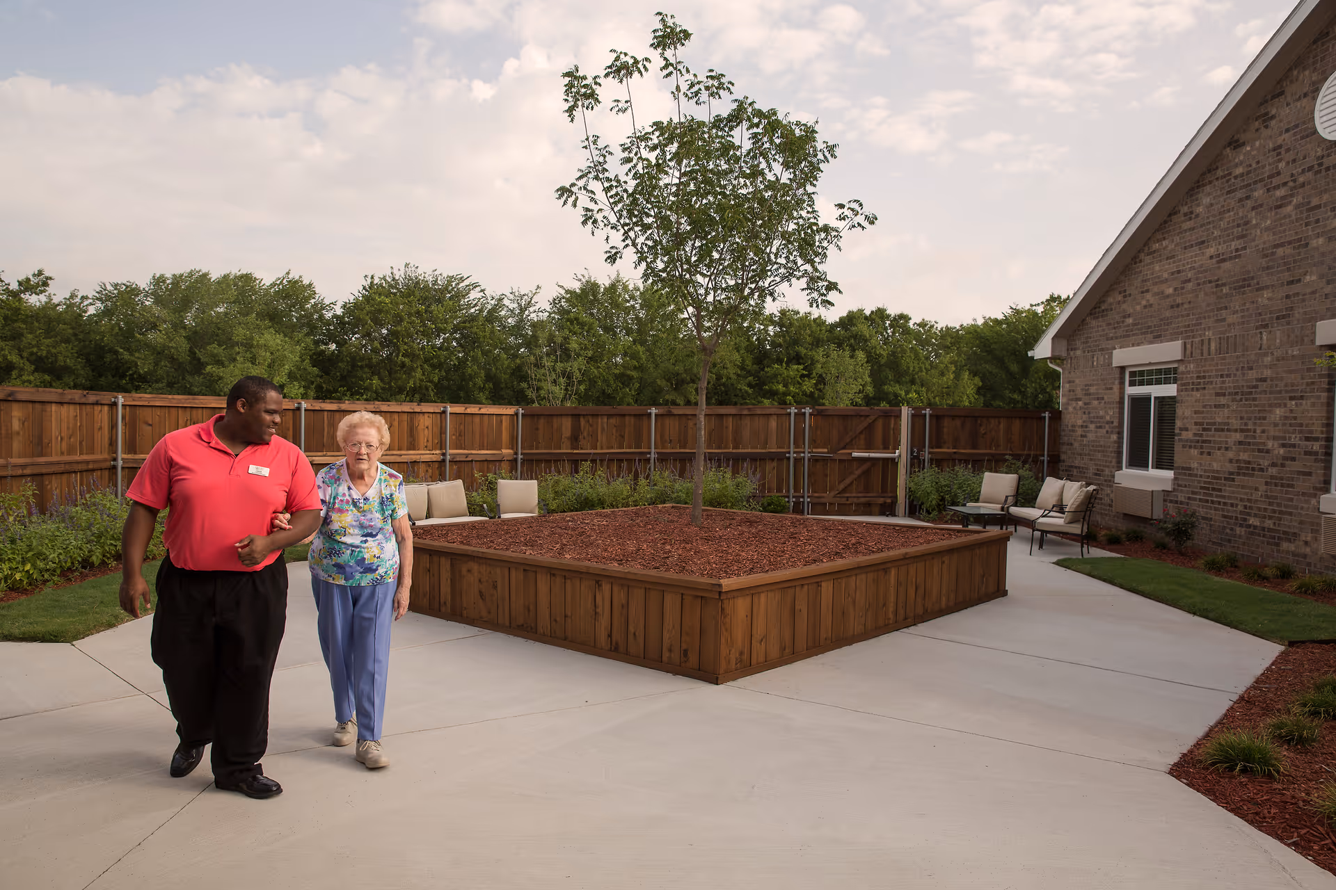 An outdoor courtyard area with a large raised wooden planter containing a small tree in the center. A man in a red shirt is walking arm-in-arm with an elderly woman wearing a floral top and blue pants. There are chairs along the wooden fence and near the brick building on the right side. The sky is partly cloudy.