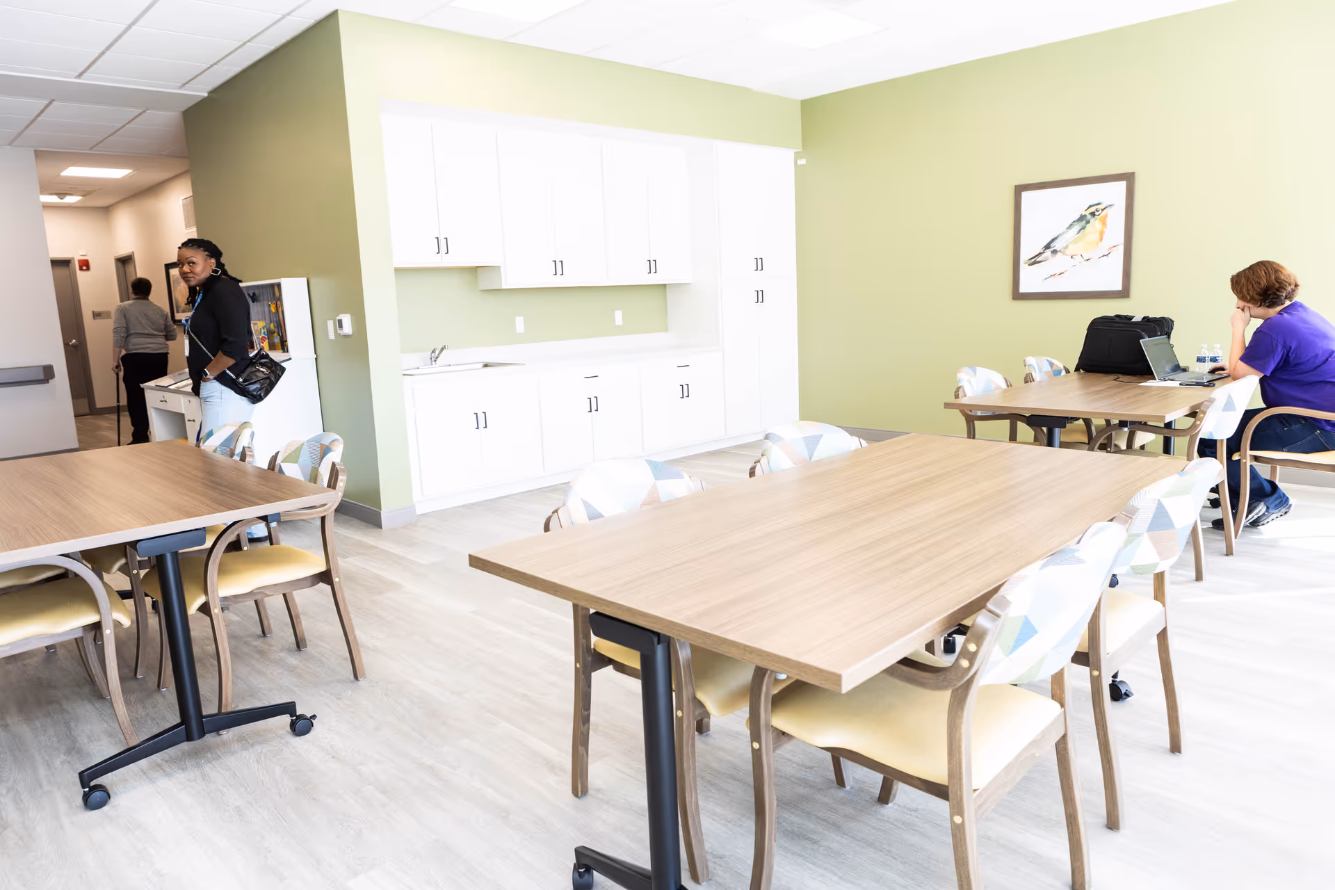 A bright room with light green walls and light wood flooring featuring two wooden tables with patterned chairs around them. A woman in a purple shirt is sitting at one table working on a laptop with water bottles nearby. White cabinets with a sink are mounted on the far wall. Two other people are visible in the background near a hallway.