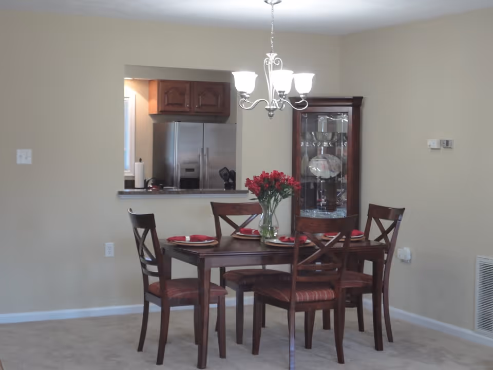 A dining area with a wooden table set for four with red napkins and a vase of red flowers in the center. Behind the table is a glass-fronted wooden cabinet displaying dishes. A kitchen with stainless steel refrigerator and wooden cabinets is visible through a pass-through window. A chandelier with five lights hangs above the table.