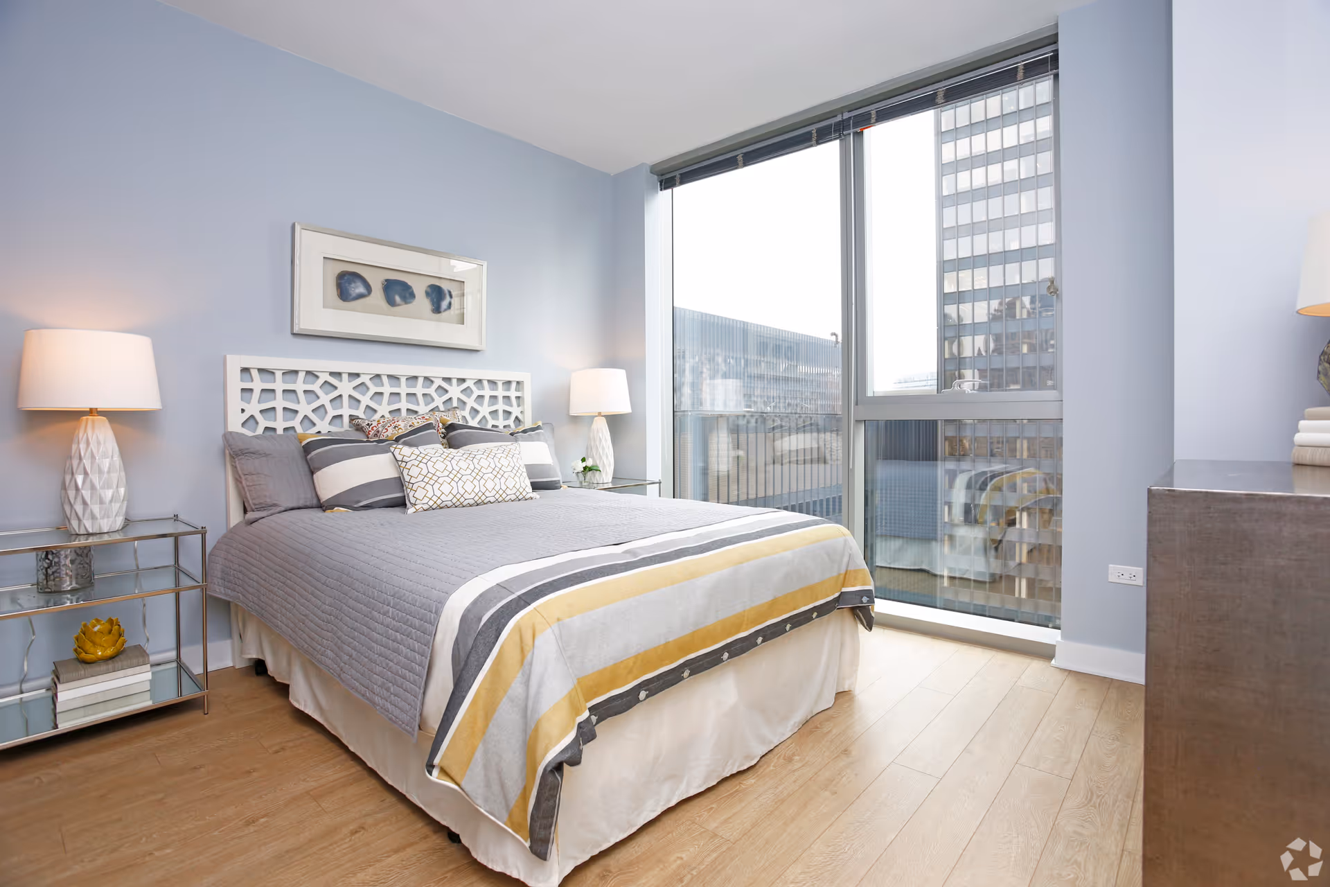 A modern bedroom with a large window showing a cityscape view. The room features a bed with a gray, white, and yellow striped comforter, multiple pillows, two bedside tables with white lamps, light blue walls, wooden flooring, and a framed artwork above the bed.