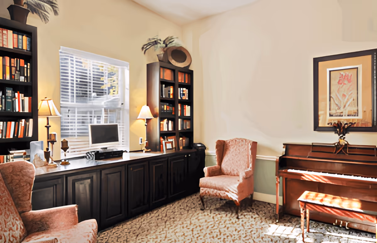 Sunlit common room with bookshelves and a desk under a window, upholstered armchairs, and an upright piano.
