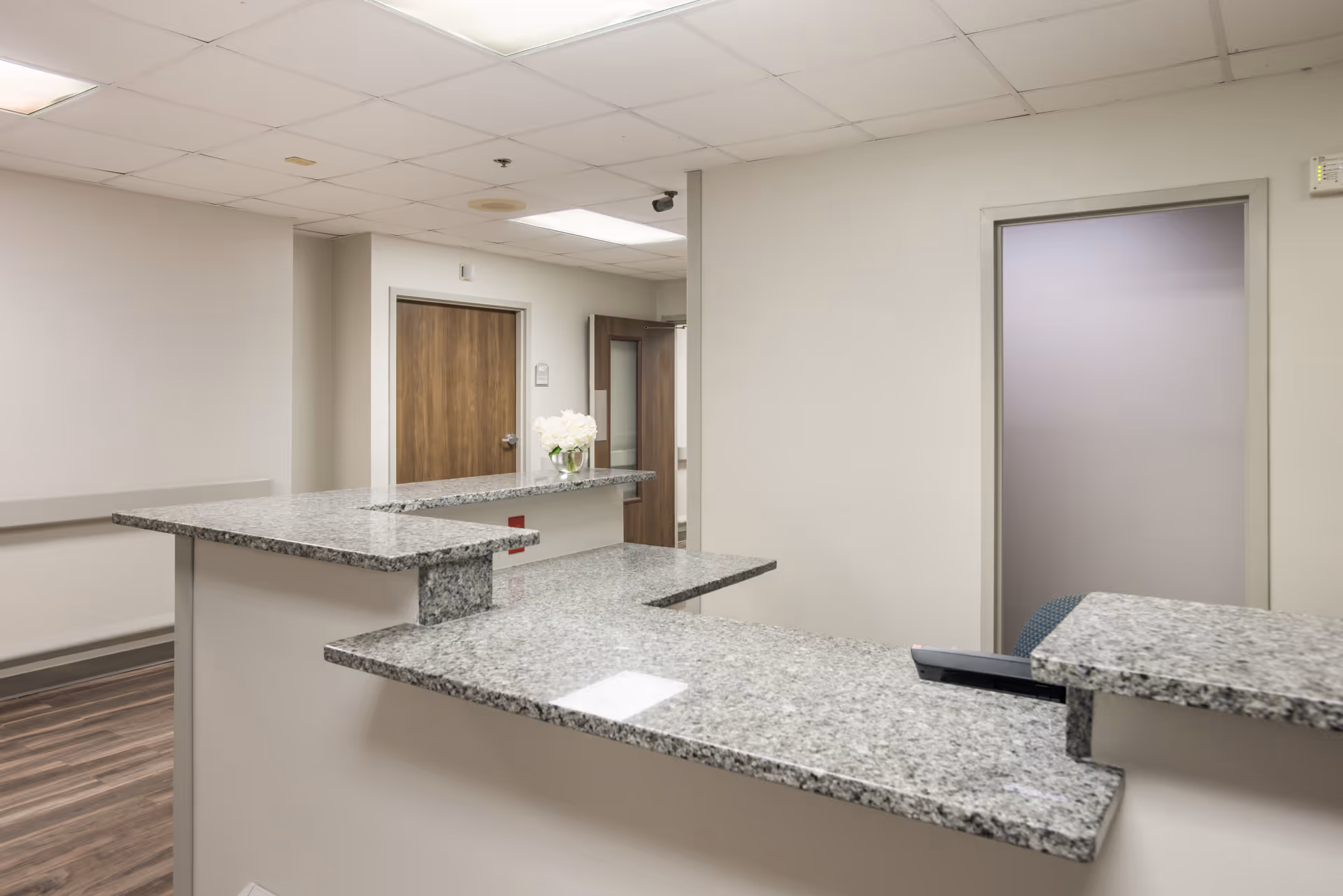 Reception area with a granite countertop desk, a vase with white flowers on the counter, wood doors, and a frosted glass door in the background. The walls are light-colored and the floor has wood-style flooring.