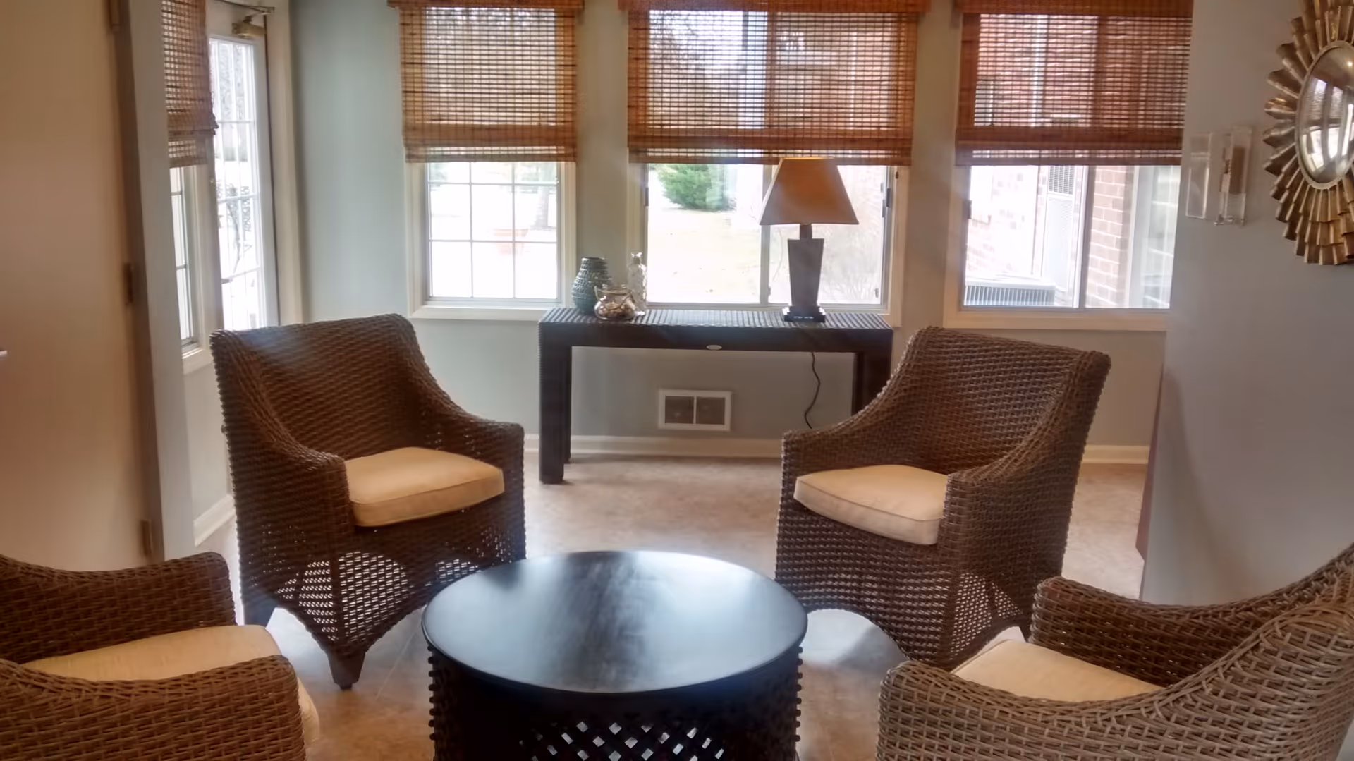 A cozy sitting area with four wicker armchairs with beige cushions arranged around a round black coffee table. Behind the chairs, there is a black console table with a lamp and decorative items placed on it. The room has large windows with bamboo blinds letting in natural light.
