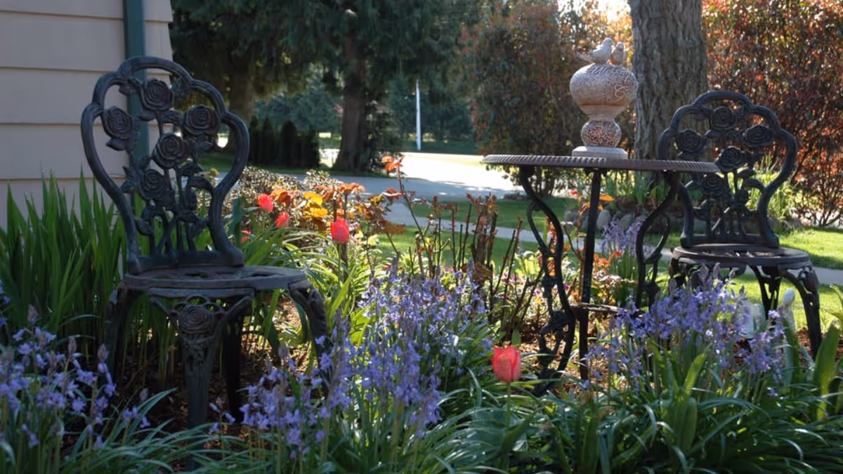 A small outdoor garden seating area with two ornate black metal chairs and a matching round table surrounded by blooming purple and red flowers, with trees and a pathway in the background.