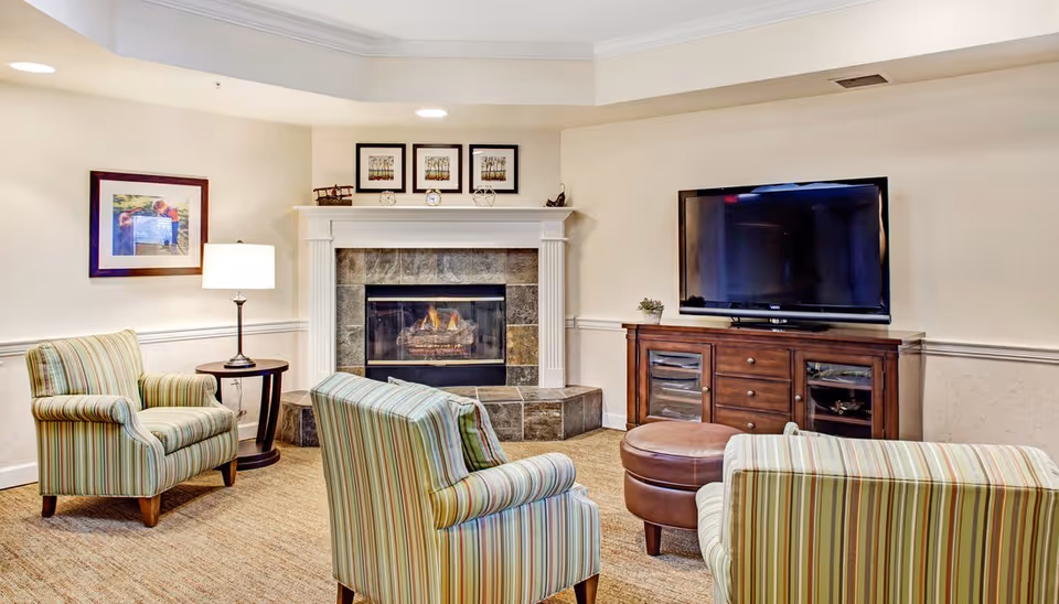Cozy communal living room with striped armchairs and an ottoman arranged around a stone fireplace, a lamp, framed art, and a TV on a wooden media cabinet.