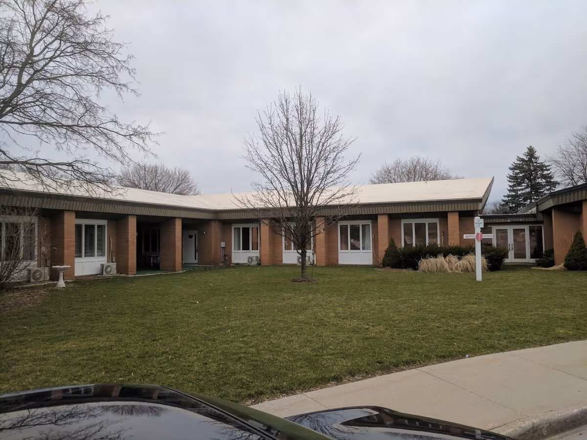 Exterior view of a single-story brick building with a flat roof, surrounded by a grassy lawn and leafless trees under a cloudy sky. There is a sidewalk in front and a signpost on the lawn.