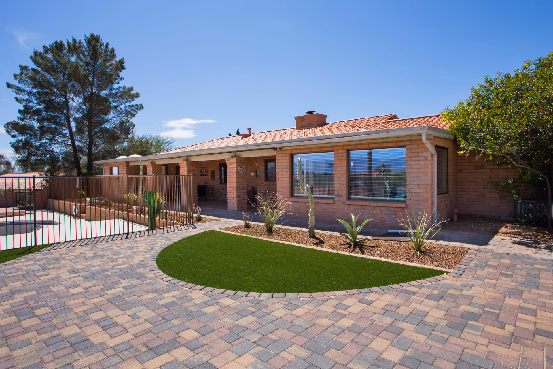 Single-story brick assisted living building with a paved courtyard, small landscaped turf area and a fenced pool under a clear blue sky.