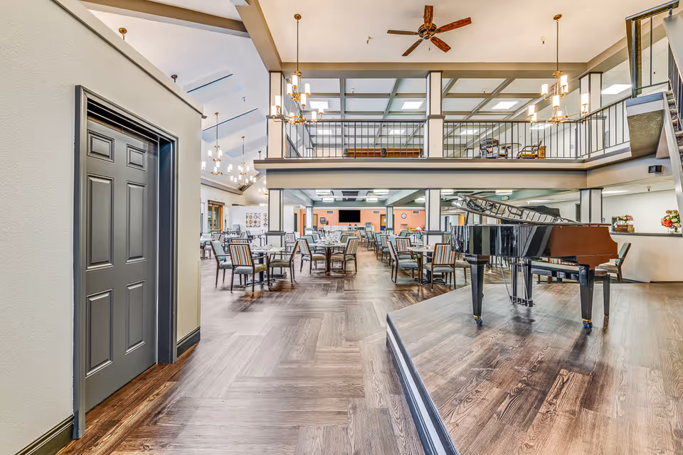 Spacious senior living dining area with multiple tables and chairs arranged neatly. A grand piano is placed on a raised platform to the right. The room features high ceilings with chandeliers and a mezzanine level with railings overlooking the dining space. The flooring is wood with a herringbone pattern, and the walls are painted in neutral tones.