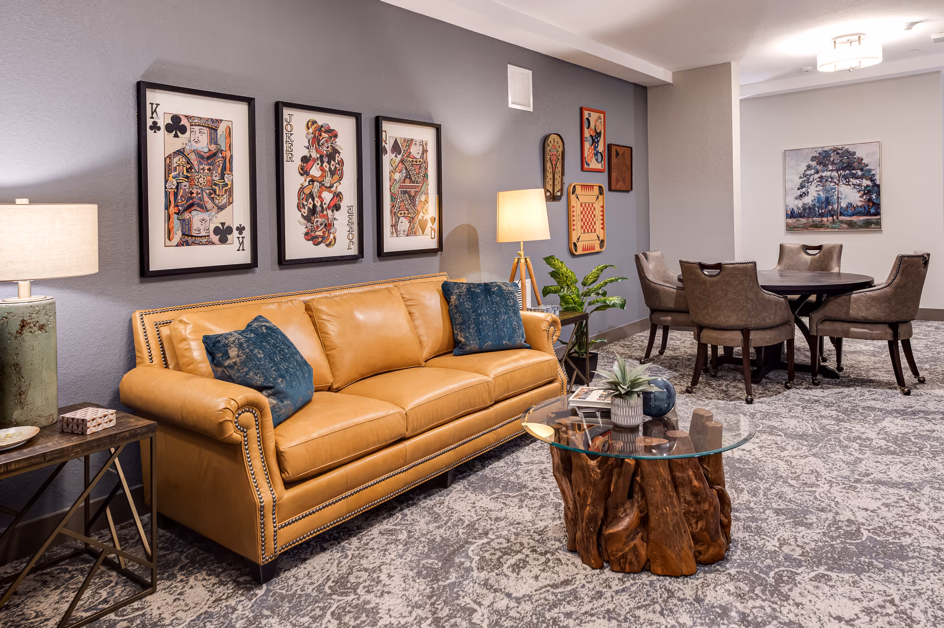 A cozy living room area featuring a tan leather sofa with two blue cushions, a glass-top coffee table with a wooden base, and a side table with a lamp. The wall behind the sofa is decorated with framed playing card artwork and various game boards. In the background, there is a round table surrounded by four upholstered chairs and a painting of a tree on the wall.
