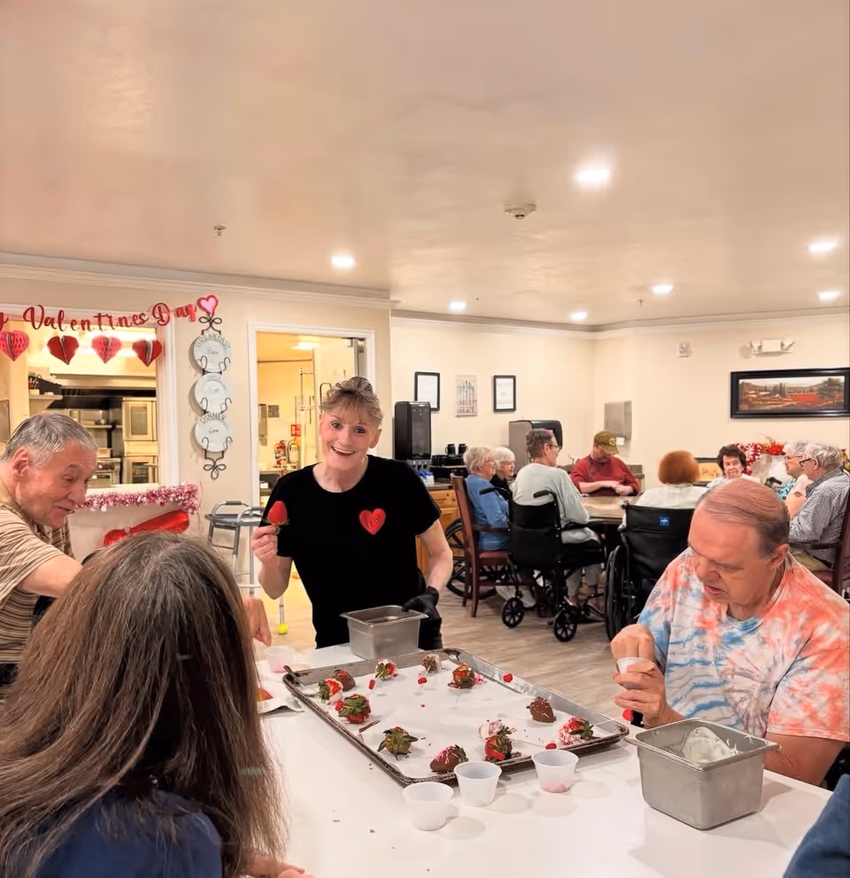 A group of senior residents and a staff member are gathered around a table in a communal dining or activity area. The staff member is smiling and holding a chocolate-covered strawberry, while the residents are engaged in decorating or eating strawberries. The room is decorated with Valentine's Day themed decorations, including hanging hearts and a sign. Other residents are seated at tables in the background, some in wheelchairs, enjoying the social setting.