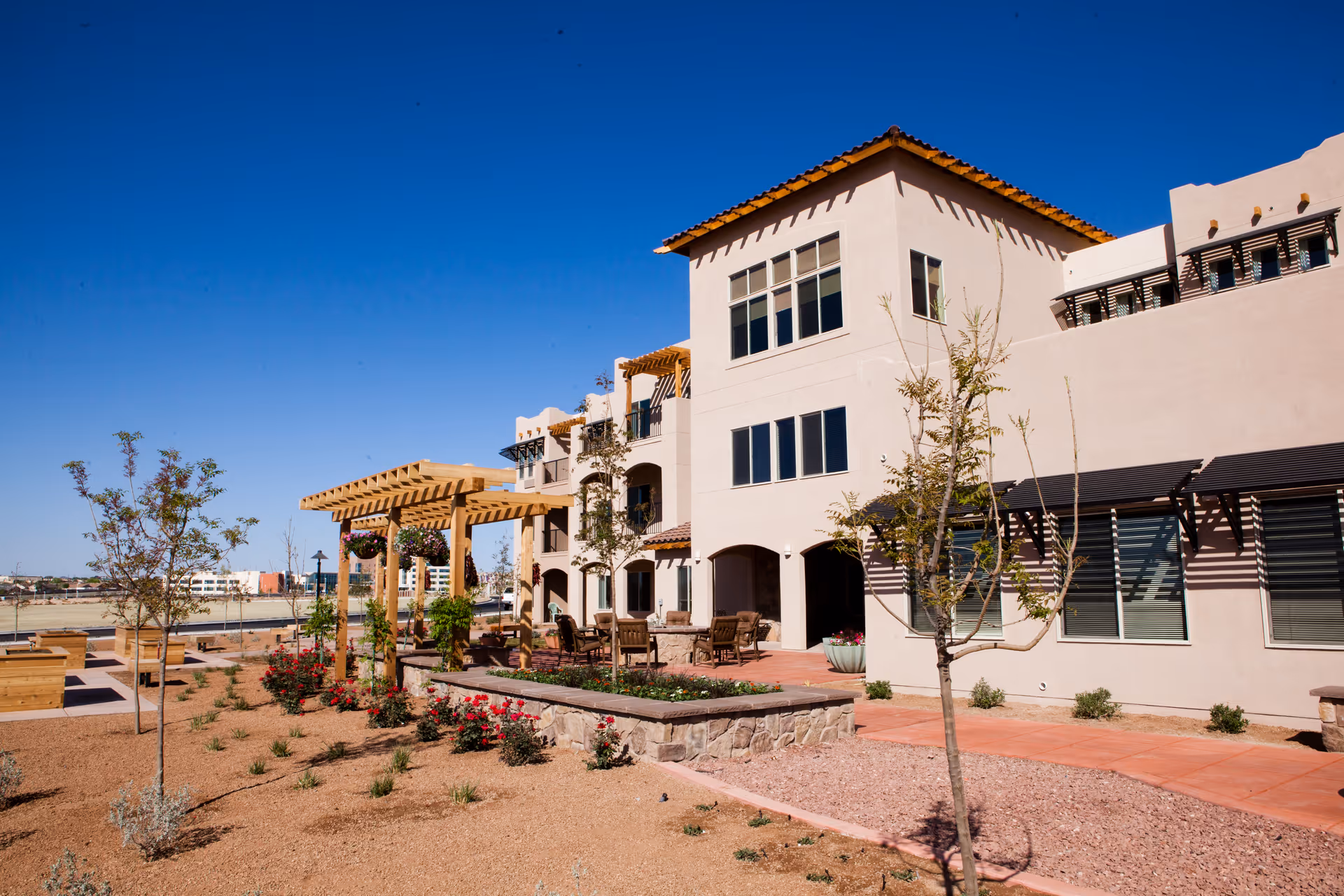 Exterior view of a multi-story retirement living facility with beige stucco walls and a red tile roof under a clear blue sky. The landscaped outdoor area features young trees, flower beds with red flowers, a wooden pergola with hanging plants, and outdoor seating with tables and chairs on a paved patio.
