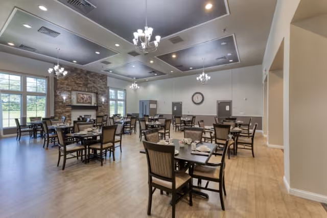 A spacious dining room with multiple tables and chairs arranged neatly. The room features large windows letting in natural light, a stone fireplace with artwork above it, chandeliers hanging from a tray ceiling, and a wall clock. The floor is wooden, and the walls are painted in neutral tones.