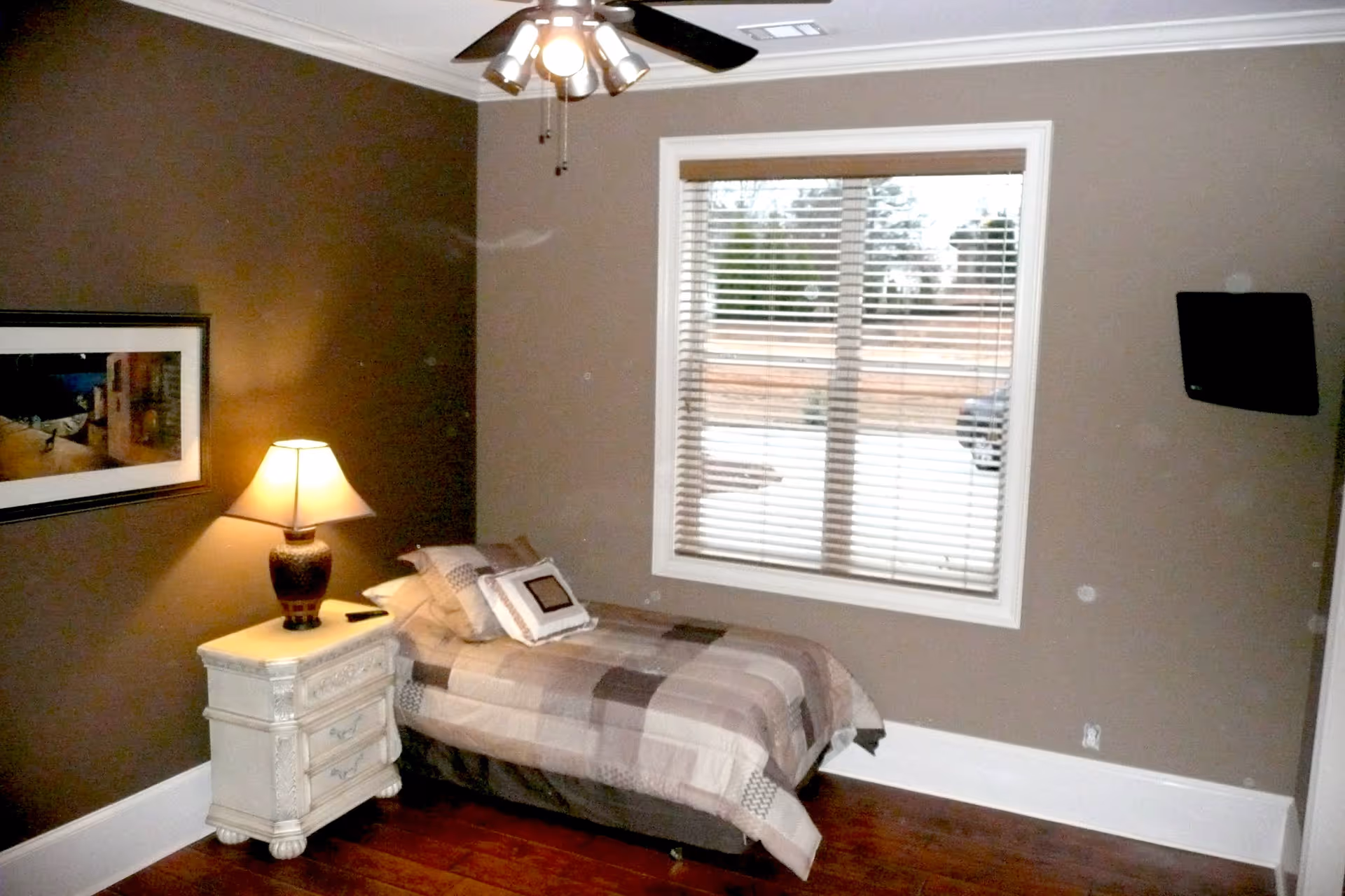 A small bedroom with a single bed covered in a patchwork quilt. Next to the bed is a white nightstand with a decorative lamp on it. A framed picture hangs on the brown wall above the nightstand. A window with white blinds is on the adjacent wall, and a ceiling fan with lights is visible above. The floor is wooden.