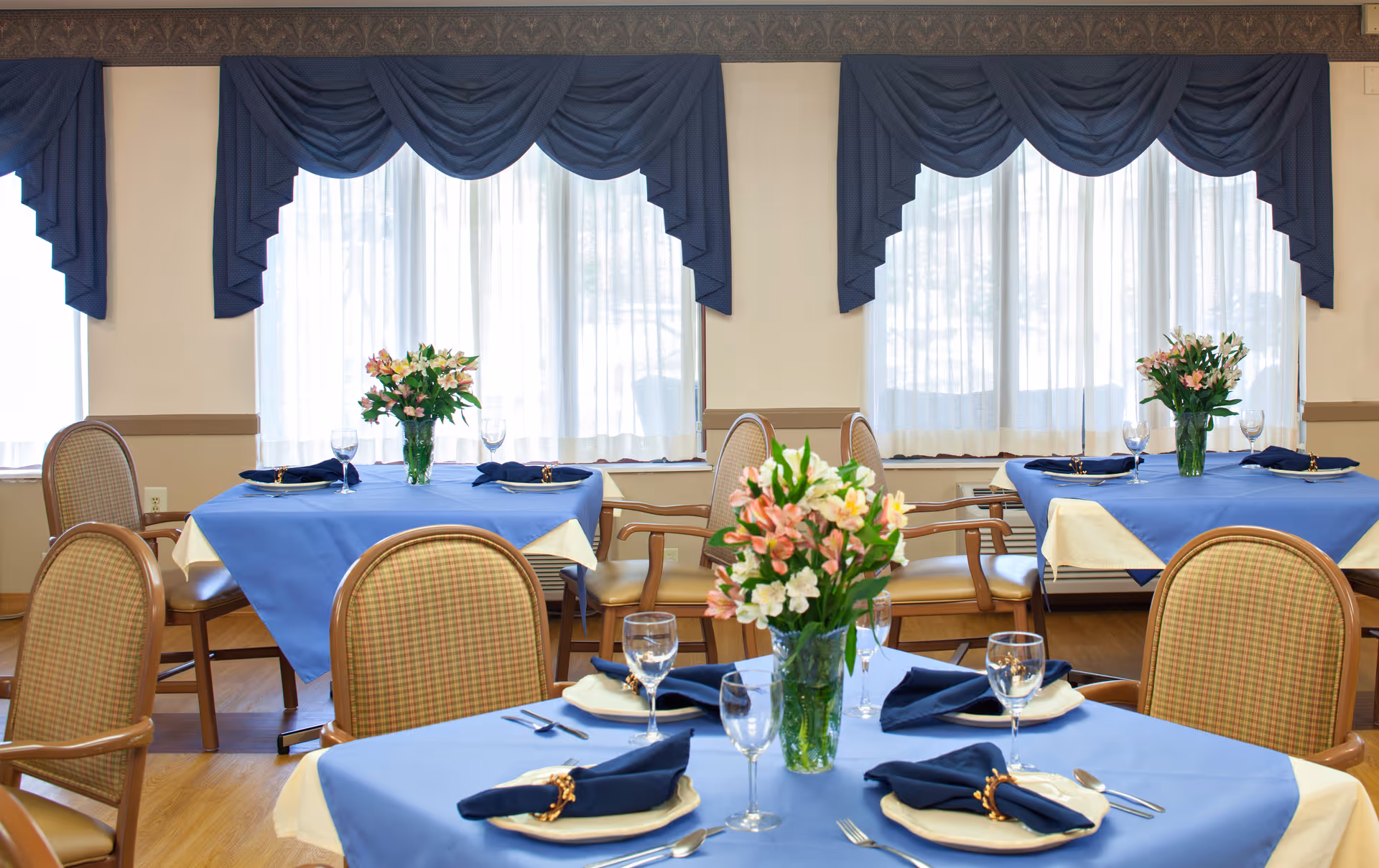 Dining room with tables covered in blue tablecloths and beige underlays, each set with plates, silverware, wine glasses, and napkins. Each table has a vase with pink and white flowers. The room has large windows with sheer white curtains and blue valances.