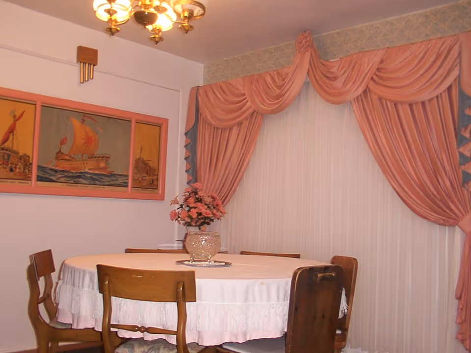 Round dining table with a floral centerpiece and wooden chairs beneath a chandelier, set against pink draped curtains and framed artwork.
