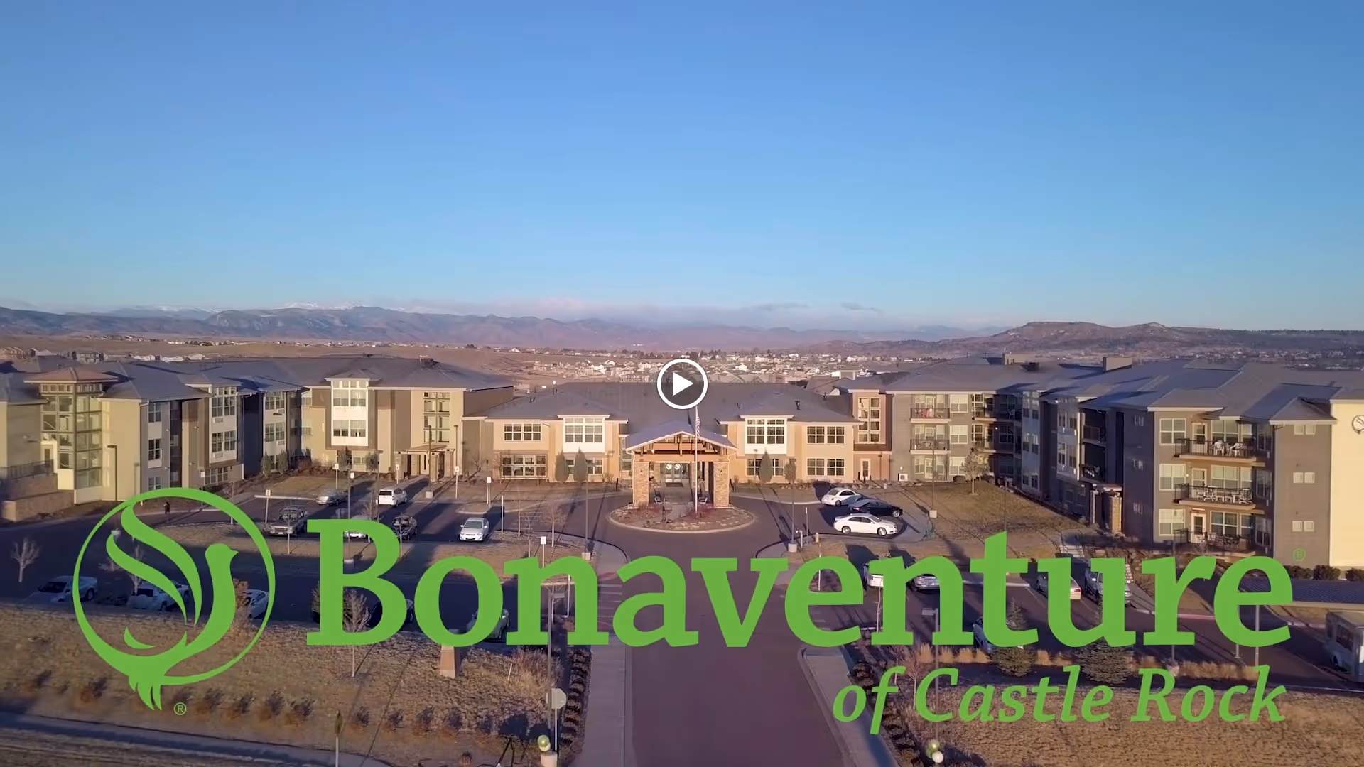 Aerial view of the Bonaventure of Castle Rock senior living facility showing multiple connected buildings with parking areas in front, set against a backdrop of mountains and a clear blue sky.