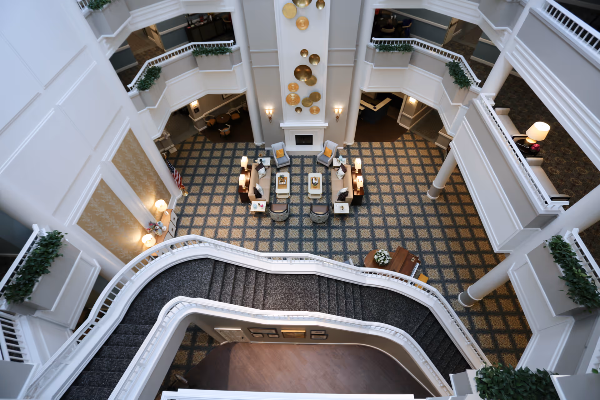 View from above of a spacious, multi-level indoor common area with a patterned carpet, a central seating arrangement with chairs and tables around a fireplace, white railings, potted plants on ledges, and decorative wall art.