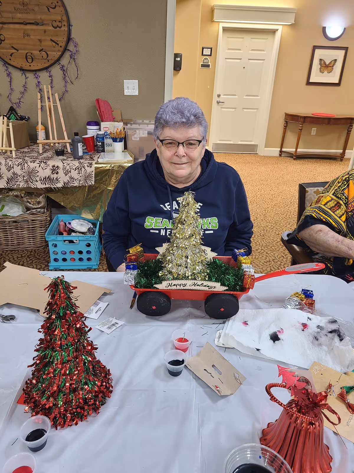 An elderly woman with short gray hair and glasses sitting at a table with holiday crafts, including a small decorated Christmas tree in a red wagon with a 'Happy Holidays' sign. The table has various craft supplies and decorations. Behind her is a wall clock, art supplies, and a door in a cozy room.