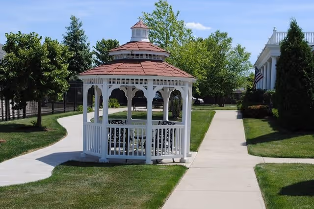 A white wooden gazebo with a red roof situated on a grassy area between two concrete walkways. Trees and shrubs surround the gazebo, and a building with white columns is visible on the right side under a clear blue sky.