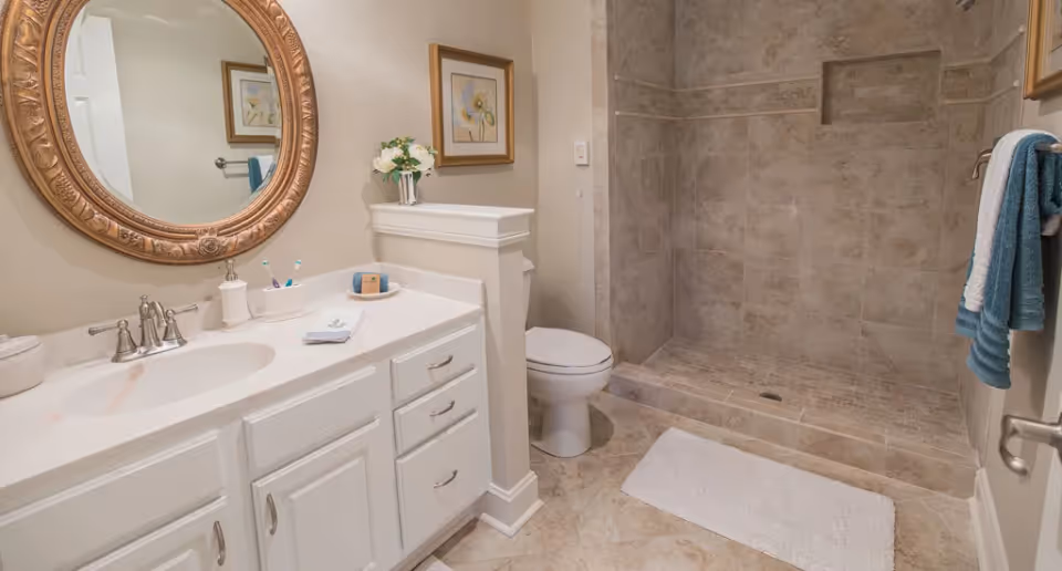 Clean white bathroom with a vanity and round gold-framed mirror, toilet, and tiled walk-in shower.