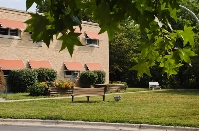 Outdoor view of a senior living facility building with beige brick walls and multiple windows with orange awnings. There are green bushes and flower beds in front of the building, a grassy lawn with two benches, and leafy tree branches partially framing the image.