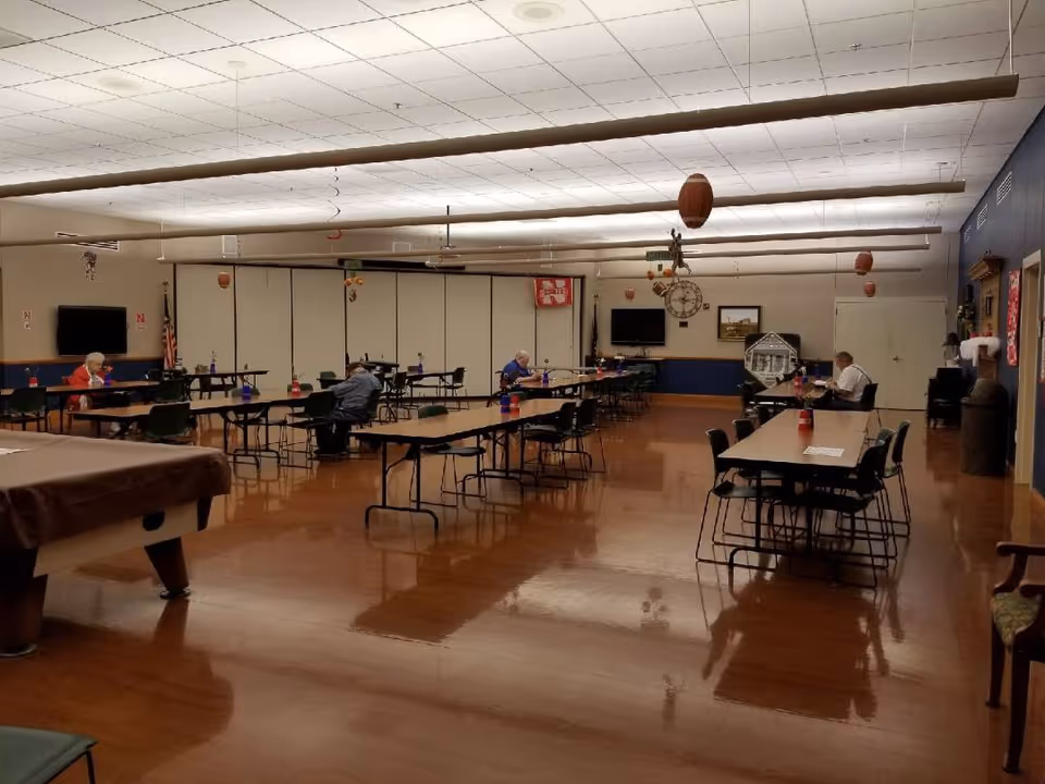 Large multipurpose dining/activity room with tables, chairs, and a few seated residents in a community facility.