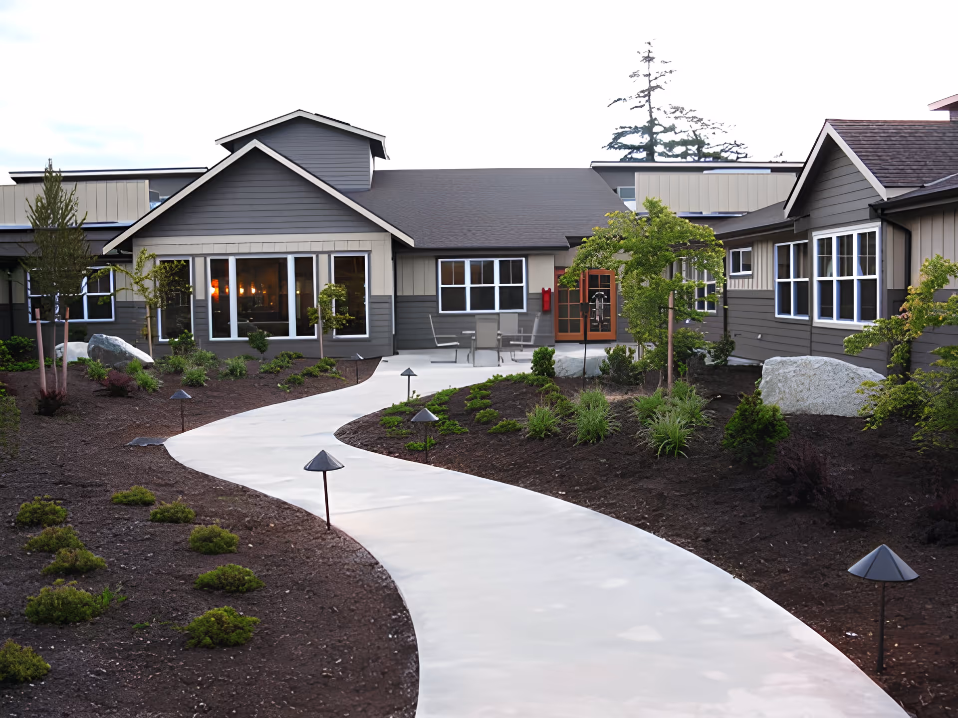 Curved concrete walkway through landscaped beds leading to the front entrance and windows of a single-story residential building.