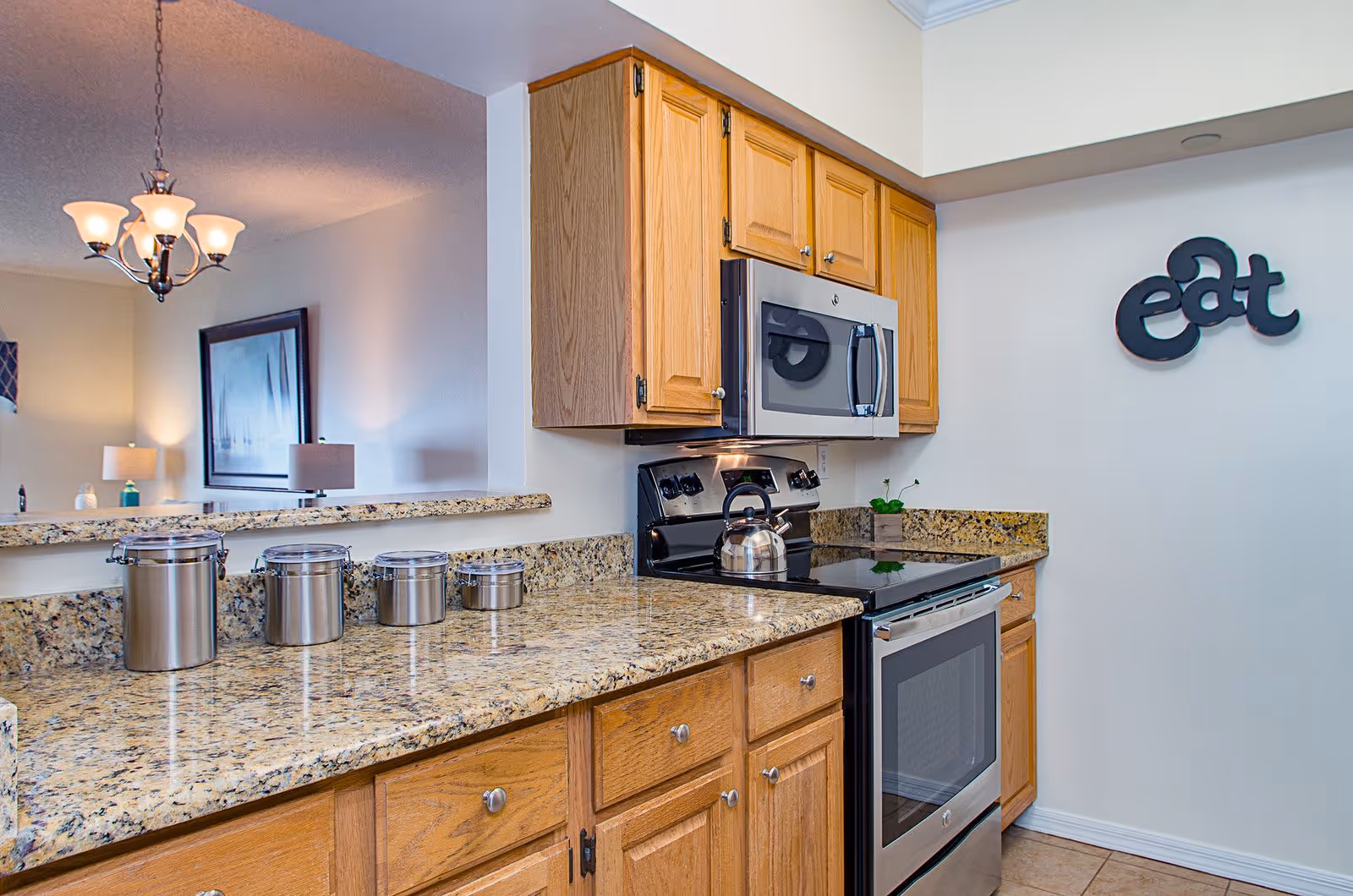 Well-lit kitchen with granite countertops, wooden cabinets, stainless steel stove and microwave, and a decorative 'eat' sign on the wall.