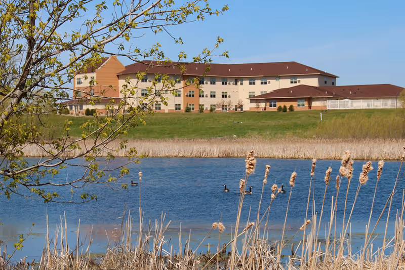 A large senior living facility building with beige and brown exterior walls and a red roof, situated behind a grassy area and a pond with reeds and ducks swimming. A tree with green leaves is visible in the foreground on the left side under a clear blue sky.