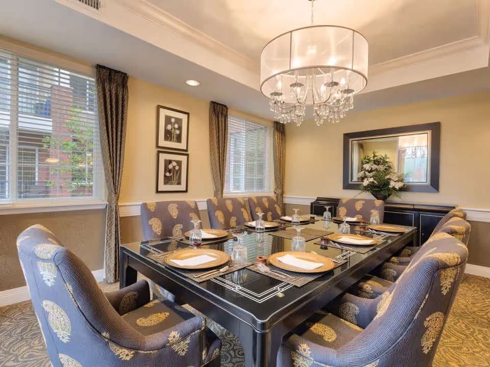 A formal dining room with a large rectangular black table set with placemats, plates, napkins, and upside-down glasses. The table is surrounded by upholstered chairs with a paisley pattern. The room has two windows with blinds and curtains, two framed floral pictures on the wall, a large mirror, a sideboard with a floral arrangement, and a chandelier with crystal accents hanging from the ceiling.